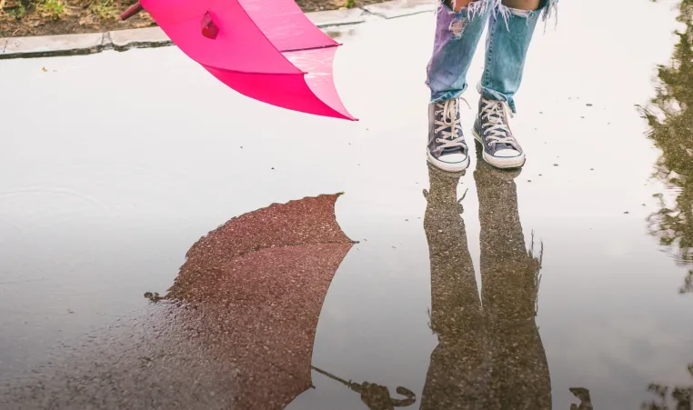 Photo of person holding a pink umbrella shown from the knees down jumping in a puddle with their reflection in the puddle.