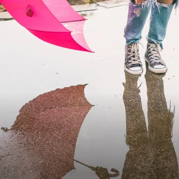 Photo of person holding a pink umbrella shown from the knees down jumping in a puddle with their reflection in the puddle.